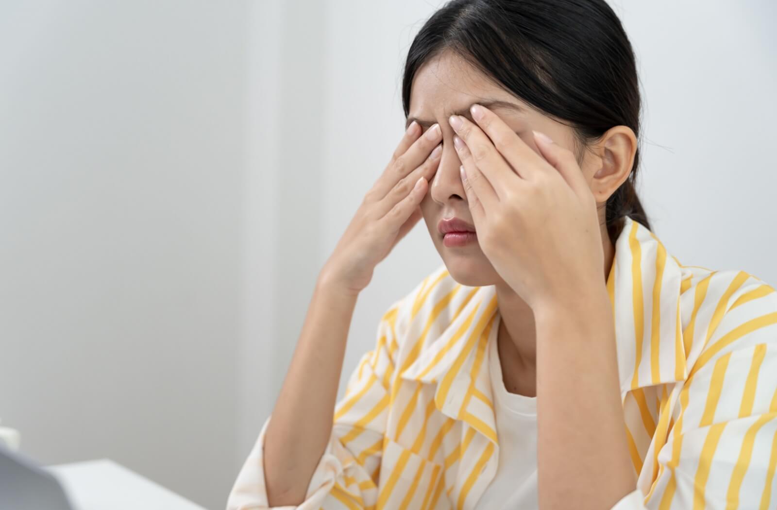 An individual in a yellow striped shirt sitting at a desk and rubbing their closed eyes in discomfort.