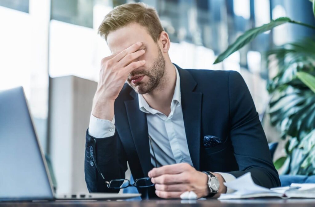 An individual in a business suit sitting at a laptop, removing their eyeglasses to rub their eyes in a bright office setting.