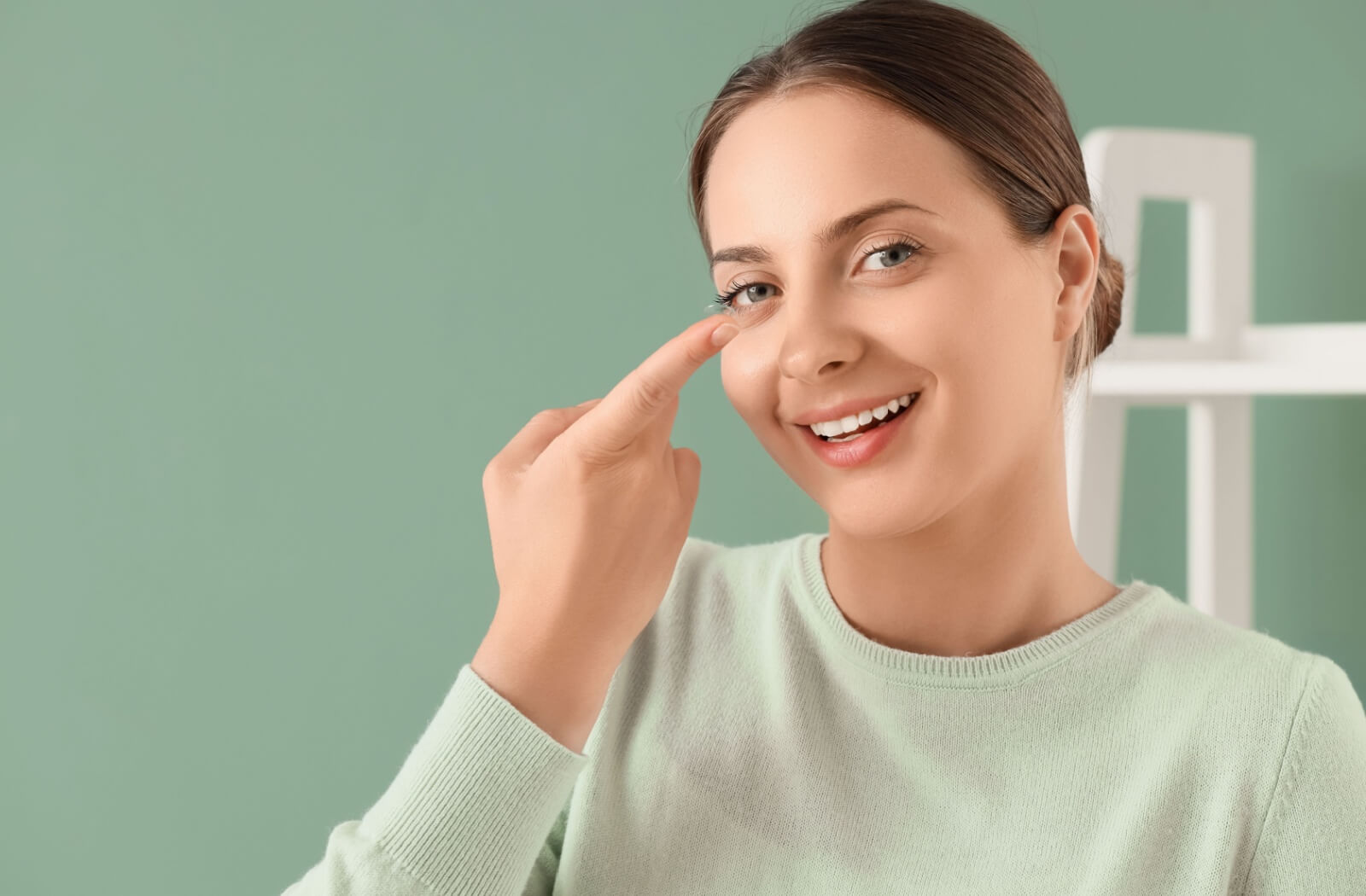 A smiling woman in a light green sweater holding a clear contact lens on her fingertip, preparing to insert it.