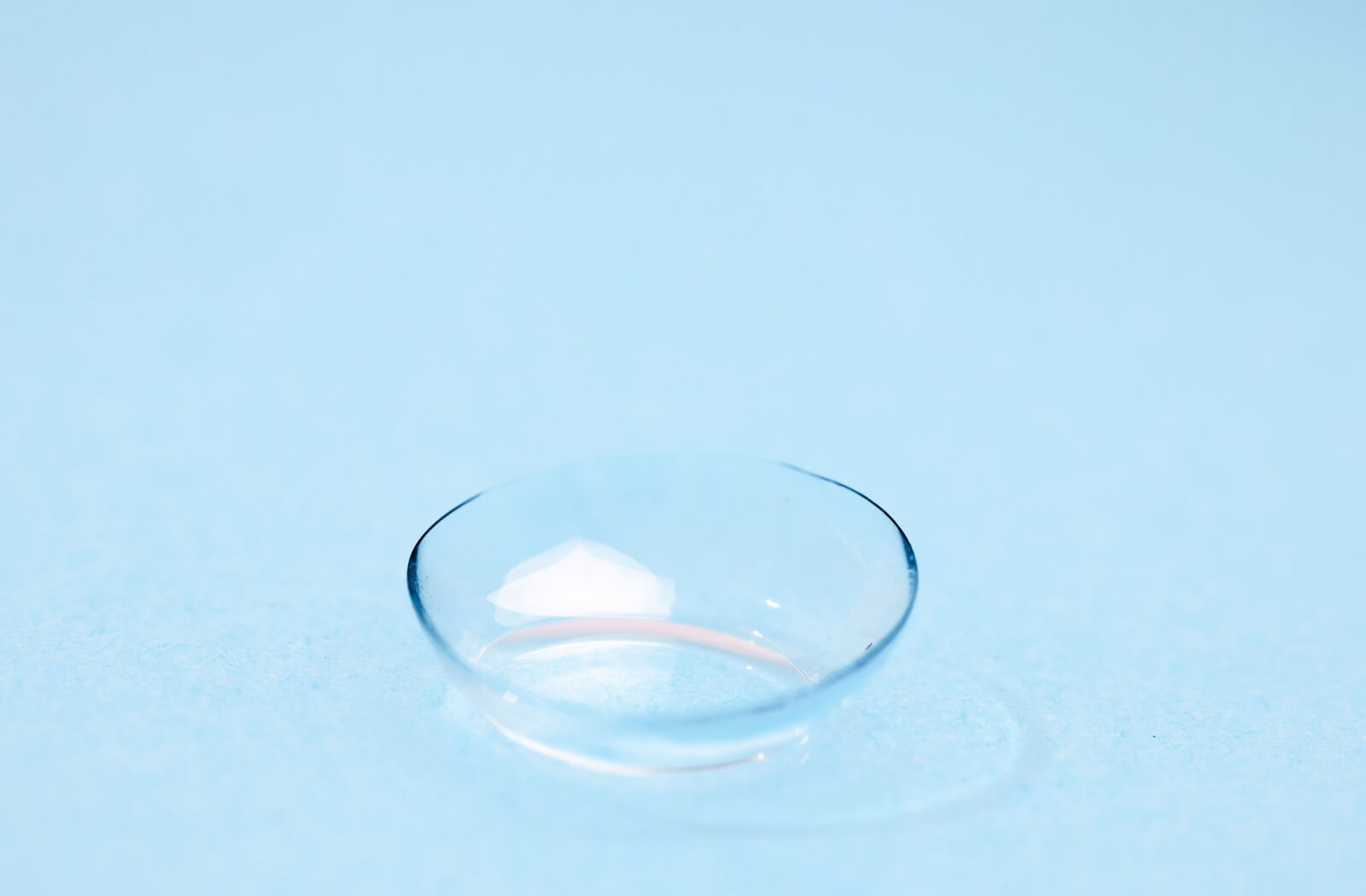 A close-up product shot of a single clear Alcon Dailies Total 1 contact lens resting upright on a smooth light blue surface.