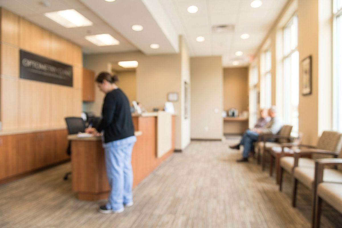 A clinic waiting room with one person standing at a reception desk and two people seated in chairs along a windowed wall.